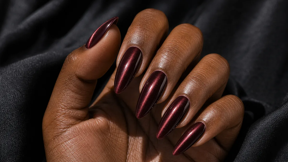Close-up of almond nails in a deep cat-eye magnetic shimmer, a moody band of light running through each nail, on deep skin on dark fabric