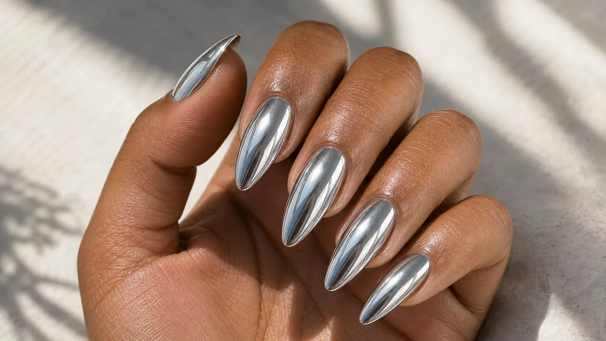 Glossy macro of almond nails in a full mirror chrome silver finish reflecting the light, the hand angled to catch the shine, on medium-deep skin