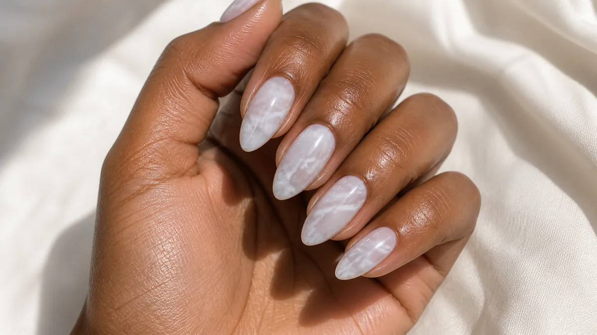 Glossy macro of almond nails in a soft grey-and-white marble swirl over a milky base, like polished stone, on deep skin catching the light