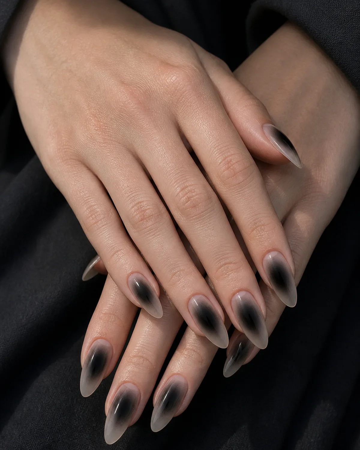 Close-up of long almond nails in a black-smoke aura, a near-transparent base with a centered black halo dissolving into smoke, on fair skin against dark fabric