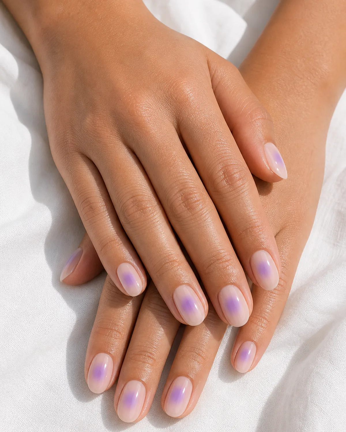 Close-up of oval nails in a lavender aura, a sheer nude base with a centered lavender halo, on medium skin resting on white fabric