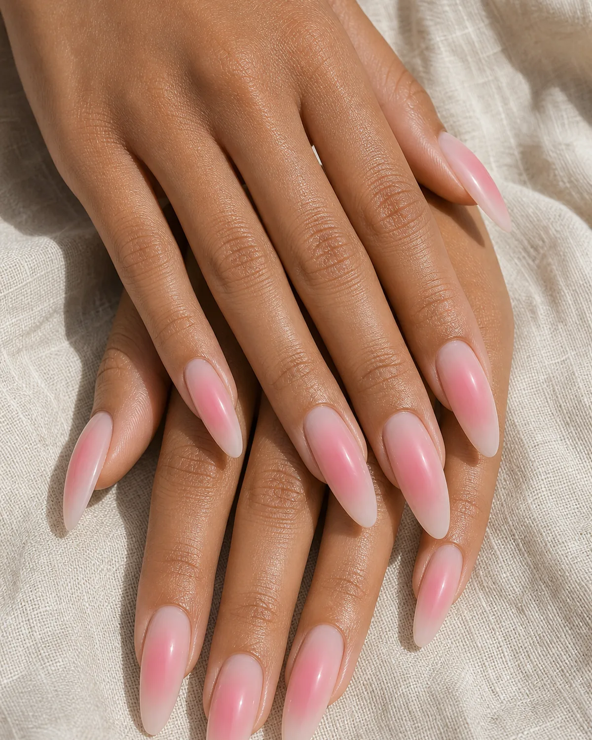 Close-up of long dramatic almond nails in a large soft-pink aura, a sheer base with a generously sized centered pink halo, on medium skin resting on oat linen