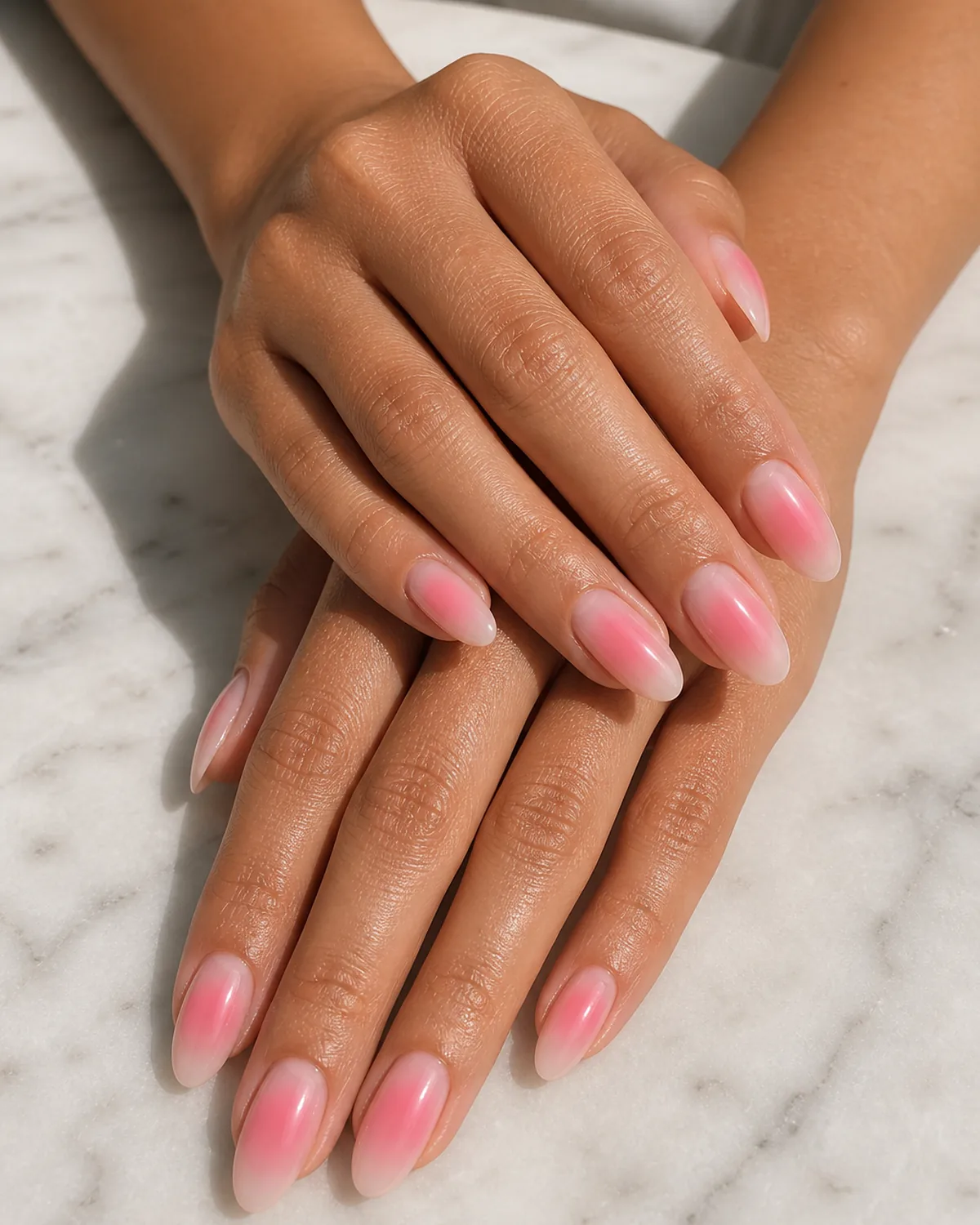 Close-up of medium almond nails in a soft pink aura, a sheer nude base with a centered pink halo, on tan skin resting on pale marble