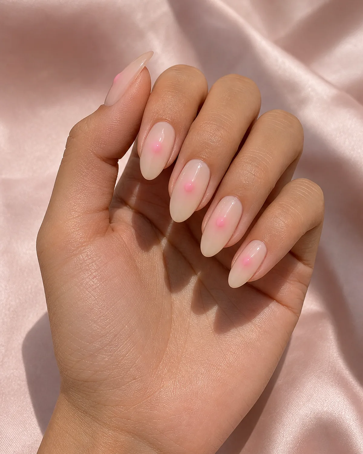 Close-up of almond nails in a single-spot center glow, one small soft pink dot centered on each nail, on light-medium skin resting on soft-pink silk