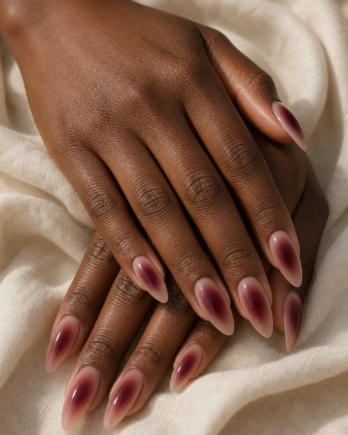 Close-up of long almond nails in a deep-wine aura, a sheer base with a velvety wine-red halo, on deep skin resting on cream fabric