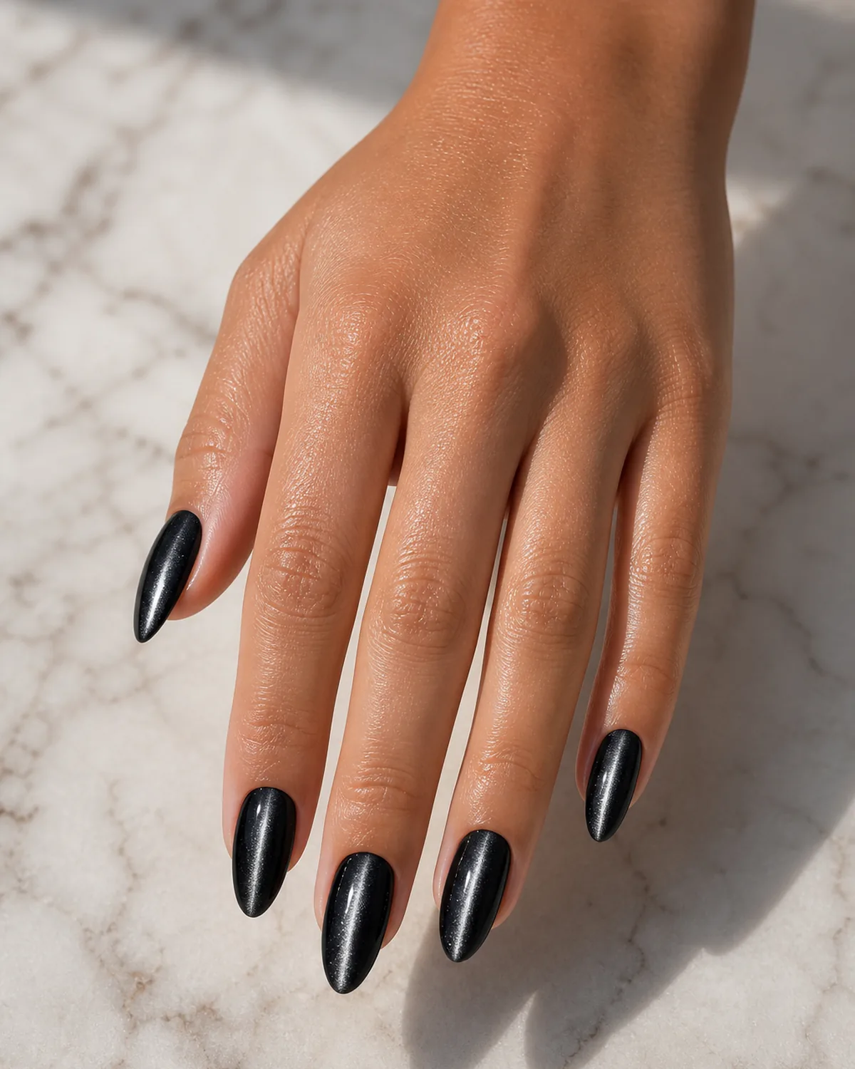 Close-up of medium almond nails in a black base with a single bright silver magnetic cat eye line running down each nail, on tan skin resting on pale marble