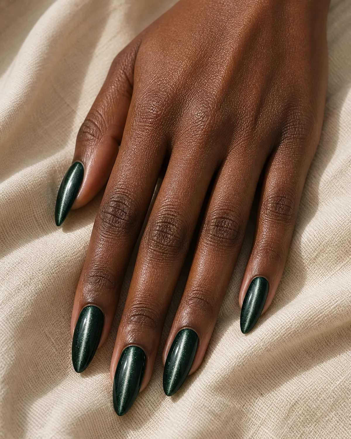 Close-up of long almond nails in a forest-green magnetic gel with a bright silver cat eye line running down each nail, on deep skin resting on cream fabric