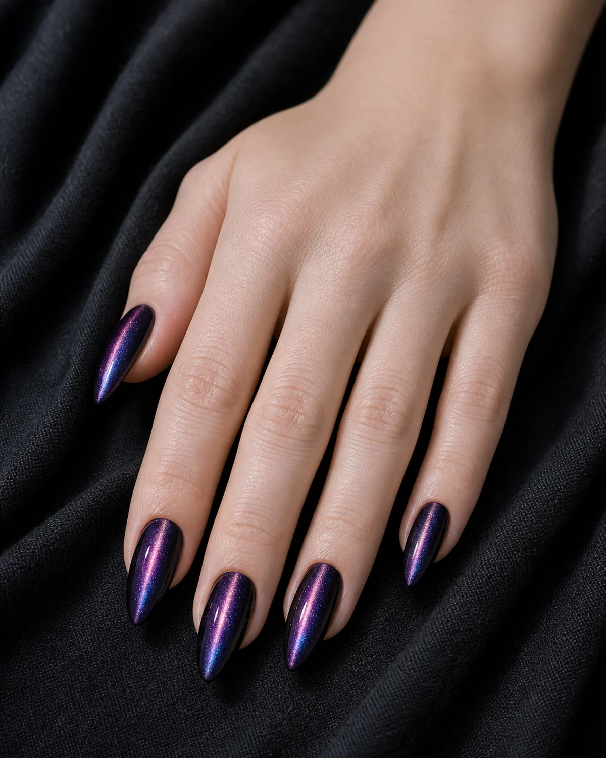 Close-up of long almond nails in a deep purple-black magnetic gel with a multi-color pink-violet-blue cat eye line on each nail, on fair skin against dark fabric