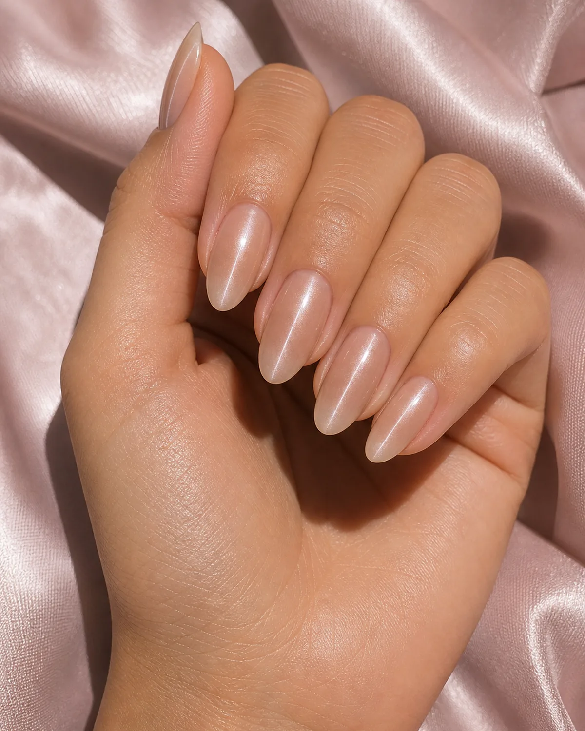Close-up of almond nails in a sheer nude magnetic gel with a thin subtle silver cat eye line on each nail, on light-medium skin resting on soft-pink silk