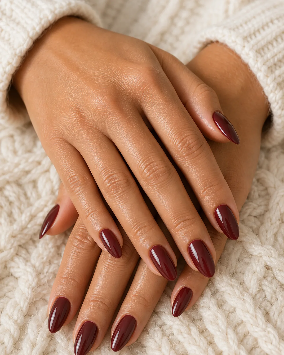 Close-up of medium almond nails in cherry mocha warm cocoa-red lacquer covering the entire nail surface from cuticle to tip, on medium skin resting on a cream knit