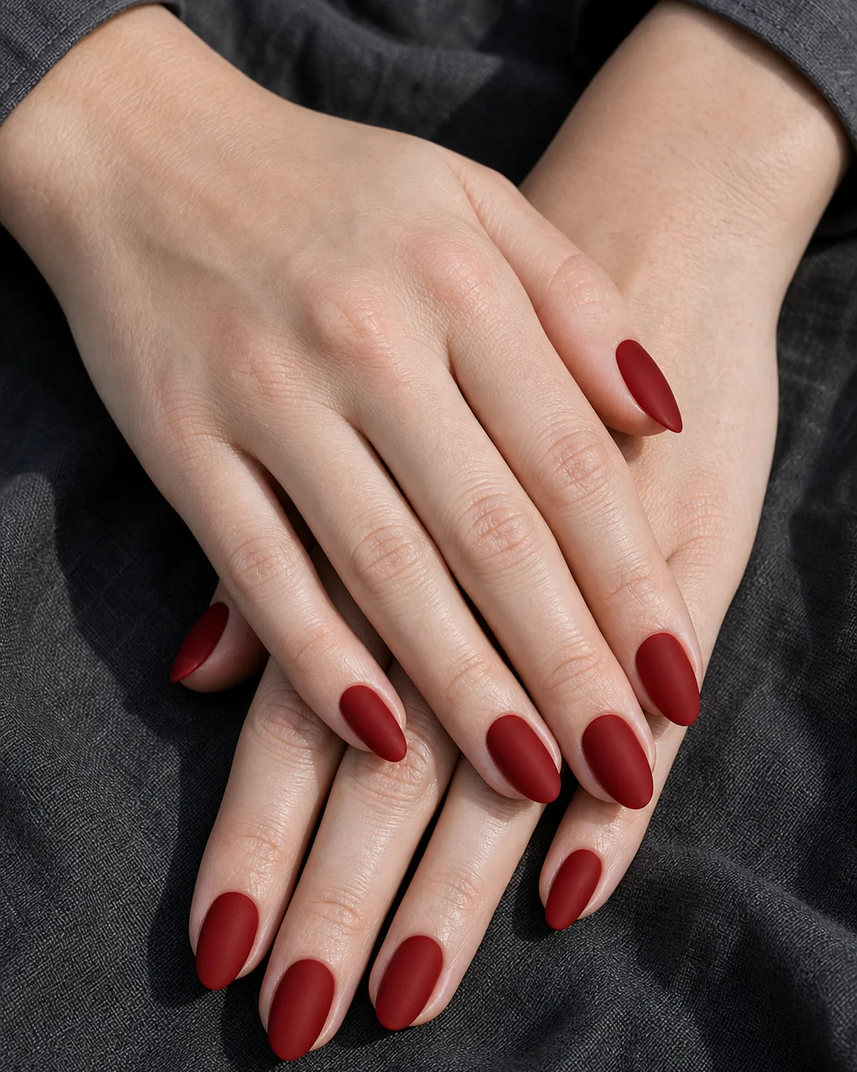 Close-up of short almond nails in matte blood-red lacquer with a flat powdery velvet finish covering the entire nail surface, on fair skin resting on charcoal linen