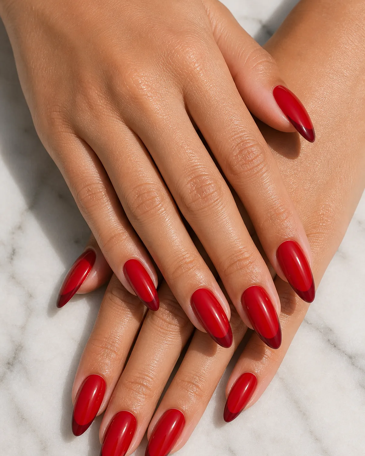 Close-up of long almond nails in red-on-red French with a cherry red base and a darker burgundy smile band at the tip, on light-medium skin resting on pale marble