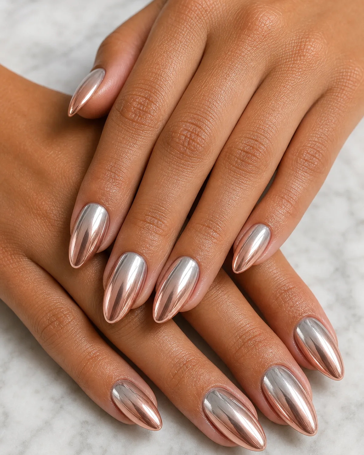 Macro of almond nails with a liquid two-tone chrome melting silver into rose gold, on medium skin against pale marble