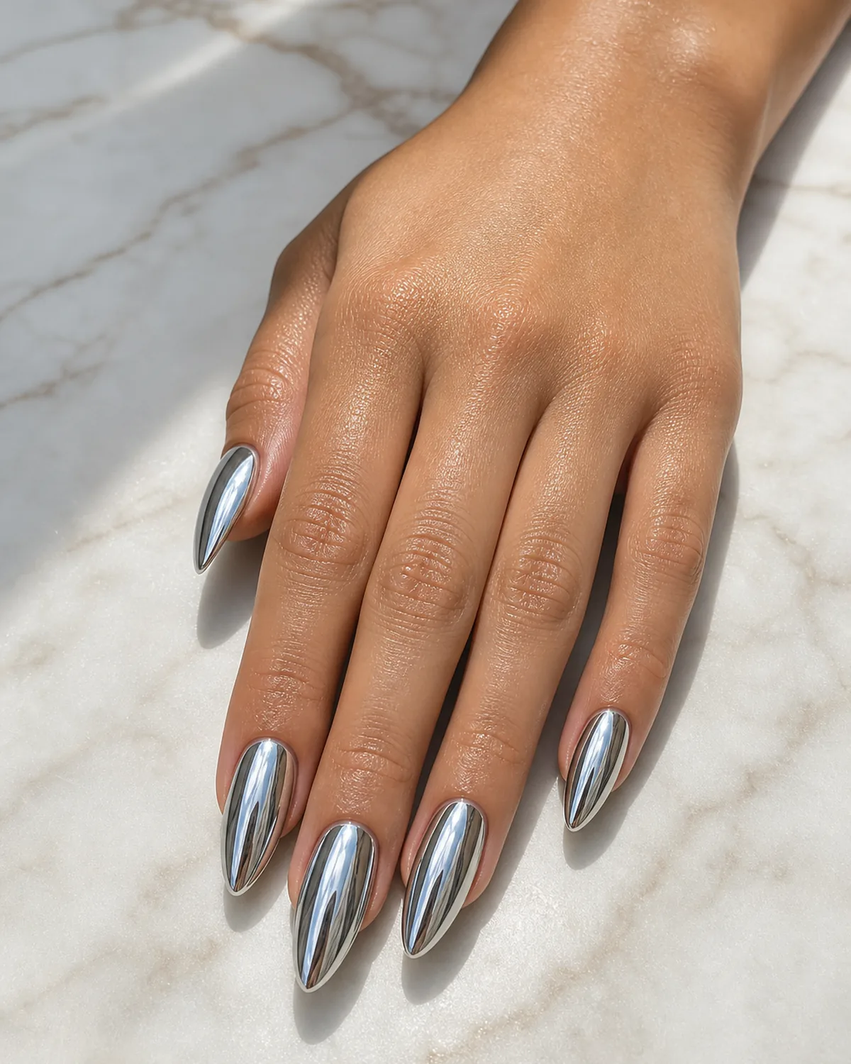 Close-up of long almond nails in a bright silver mirror chrome reflecting the light, on tan skin against pale marble