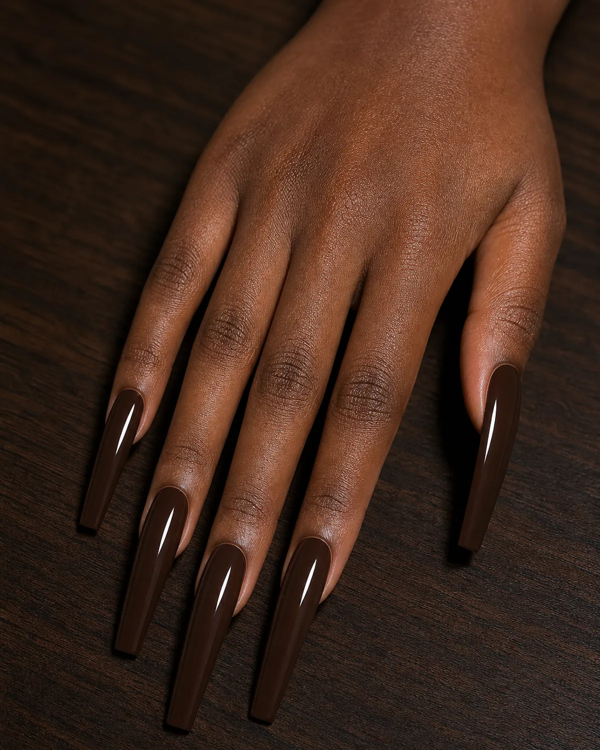 Close-up of extra-long coffin nails in rich opaque deep chocolate brown cream polish covering the entire nail with tapered side walls and a clean flat-cut squared free edge, on deep skin resting on dark walnut wood