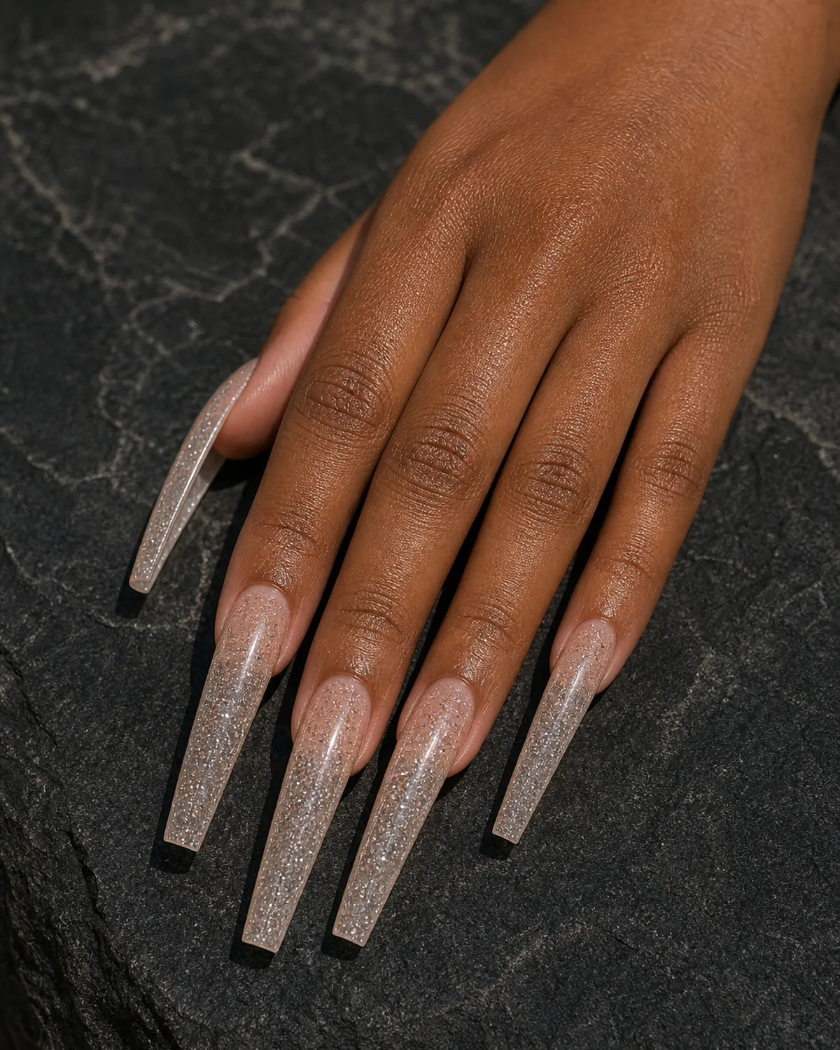 Close-up of extra-long coffin nails in a sheer clear polish suspended with fine silver glitter across the whole nail, tapered side walls and flat-cut squared free edge, on medium-deep skin resting on charcoal stone under low warm light