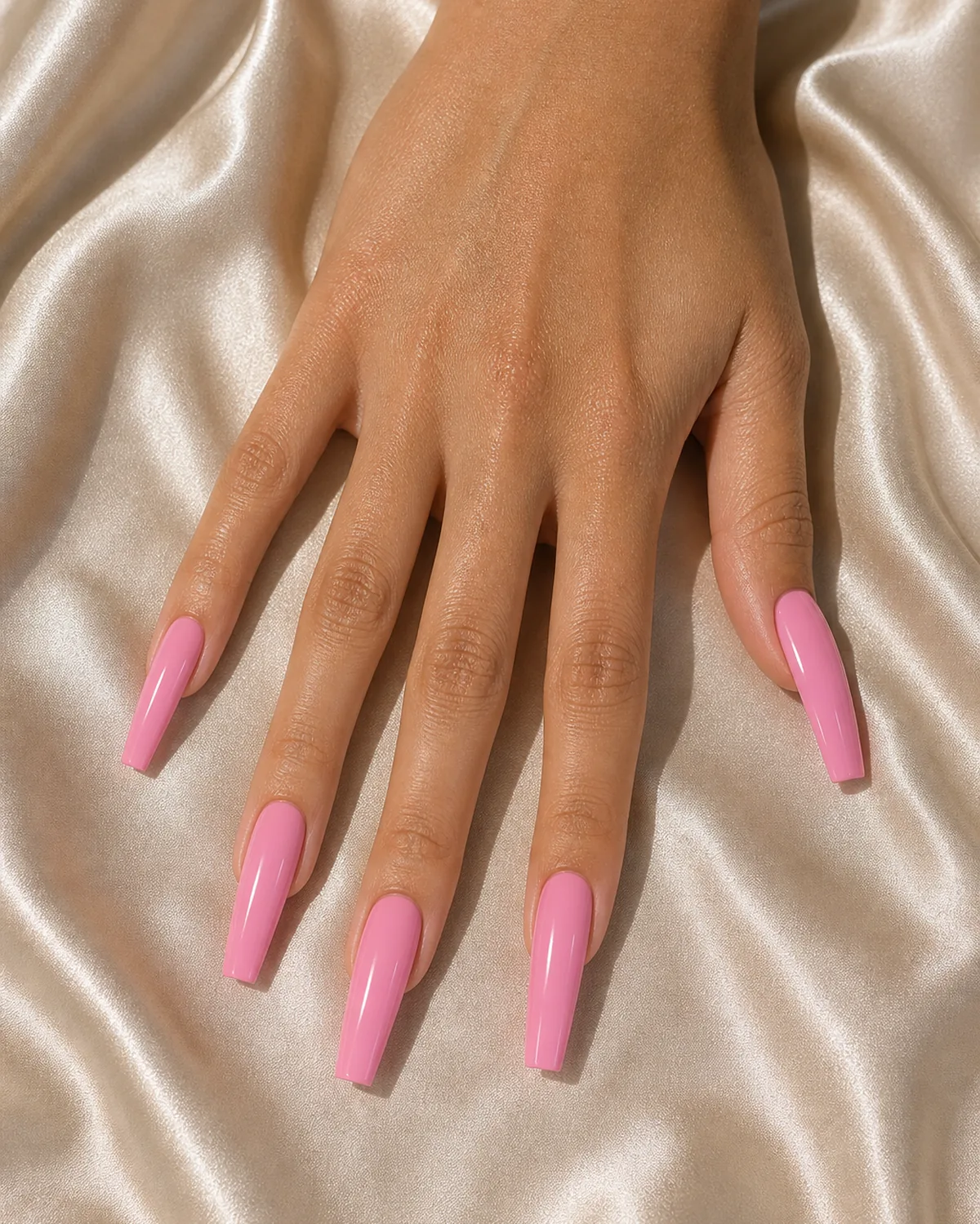 Close-up of long coffin nails in classic glossy pink polish covering the entire nail with tapered side walls and a clean flat-cut squared free edge, on medium skin resting on champagne silk