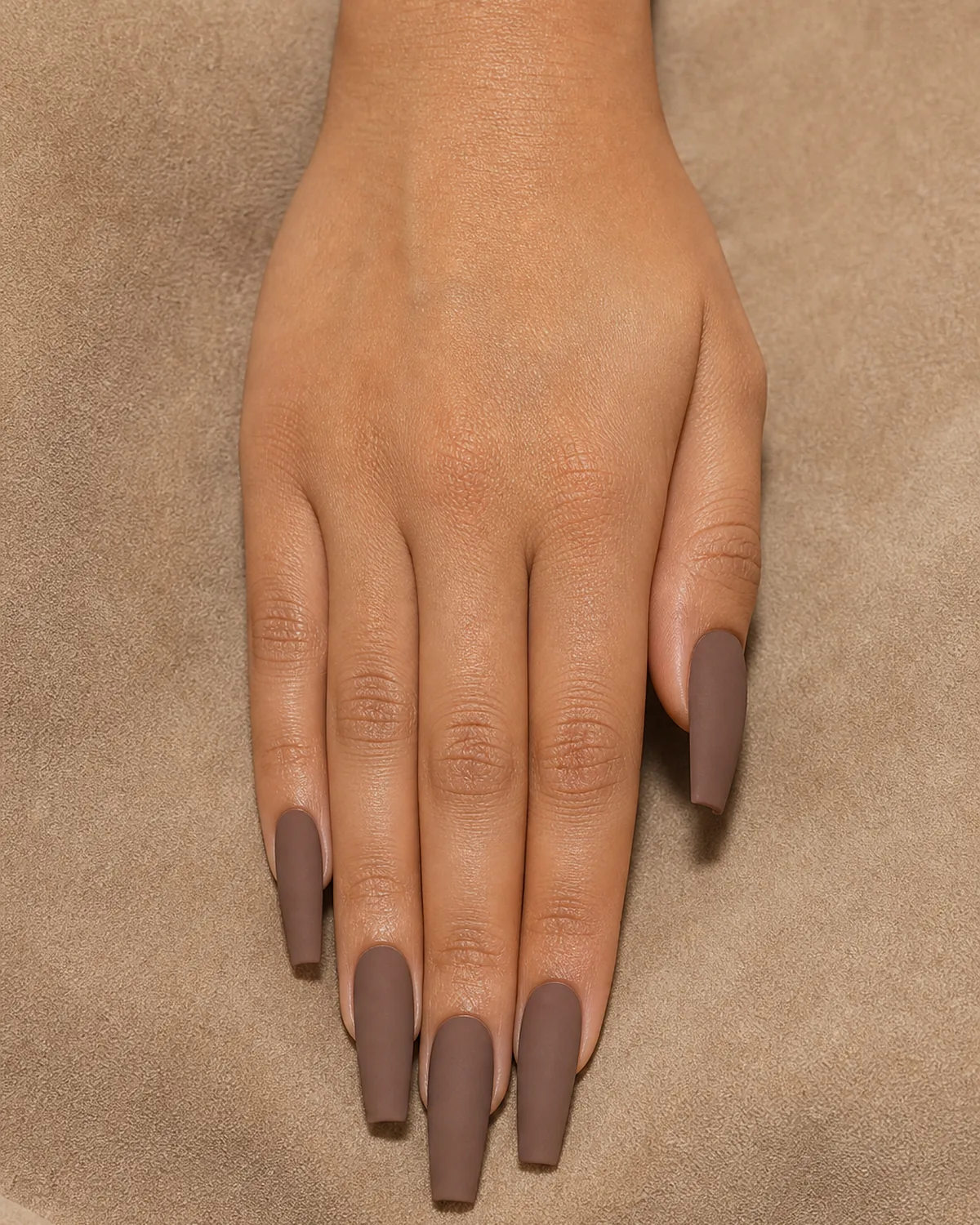 Close-up of long coffin nails in warm matte mocha polish covering the entire nail with tapered side walls and a clean flat-cut squared free edge in a flat powdery finish, on tan skin resting on warm beige suede