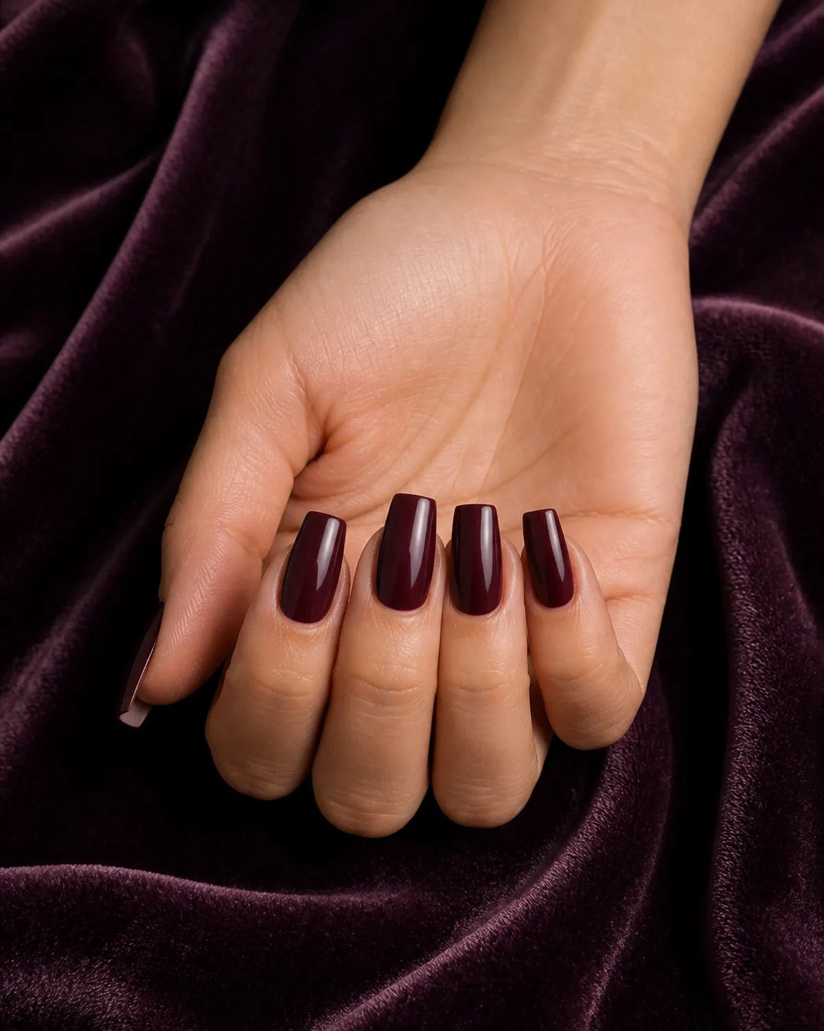 Close-up of medium coffin nails in deep burgundy wine polish covering the entire nail with tapered side walls and a flat-cut squared free edge, on light-medium skin resting on plum velvet
