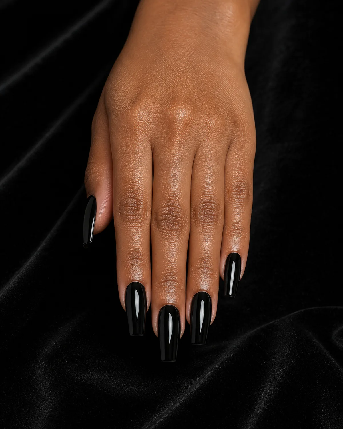 Close-up of medium coffin nails in high-gloss opaque black polish covering the entire nail with tapered side walls and a flat-cut squared free edge, on medium-deep skin resting on black velvet