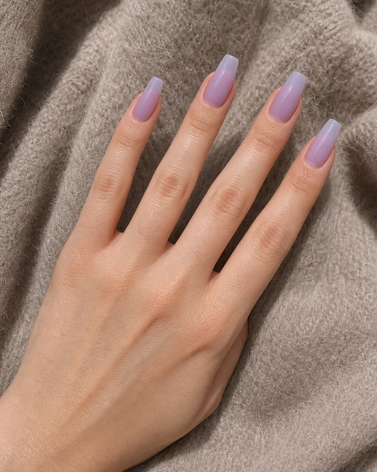 Close-up of medium coffin nails in semi-sheer cool lavender-nude polish covering the entire nail with tapered side walls and a flat-cut squared free edge, on light skin resting on soft taupe wool under soft daylight