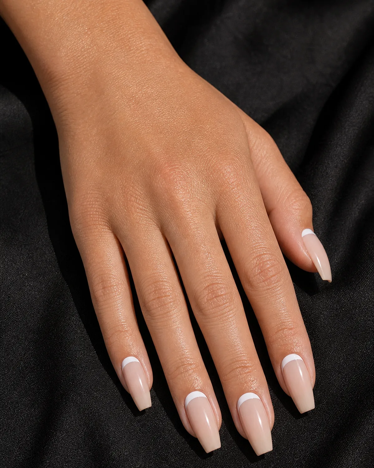 Close-up of medium coffin nails in reverse French with a clean opaque white half-moon band at the cuticle on a sheer nude base, on light-medium skin against dark fabric