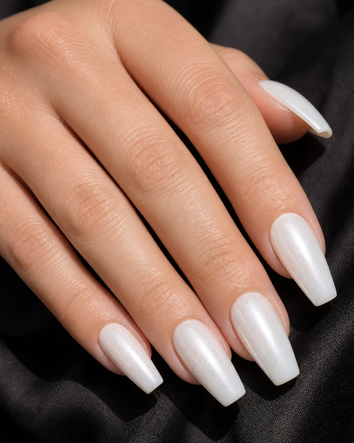 Close-up of coffin-shaped nails in a milky glazed donut finish with an architectural pearl wash, on fair skin against dark fabric