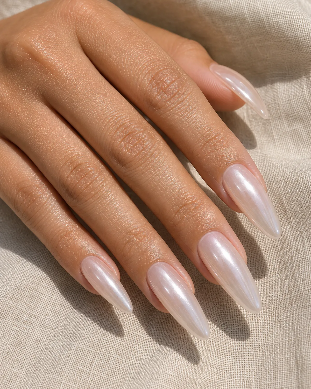 Close-up of long dramatic almond nails in a clear glazed donut finish with the deepest wet pearly sheen across the whole nail, on medium skin resting on oat linen