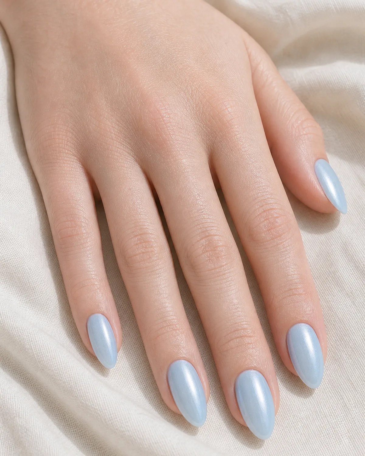 Close-up of almond nails in a sky-blue glazed donut finish with an icy pearl chrome-dust topper, on fair skin resting on cream fabric
