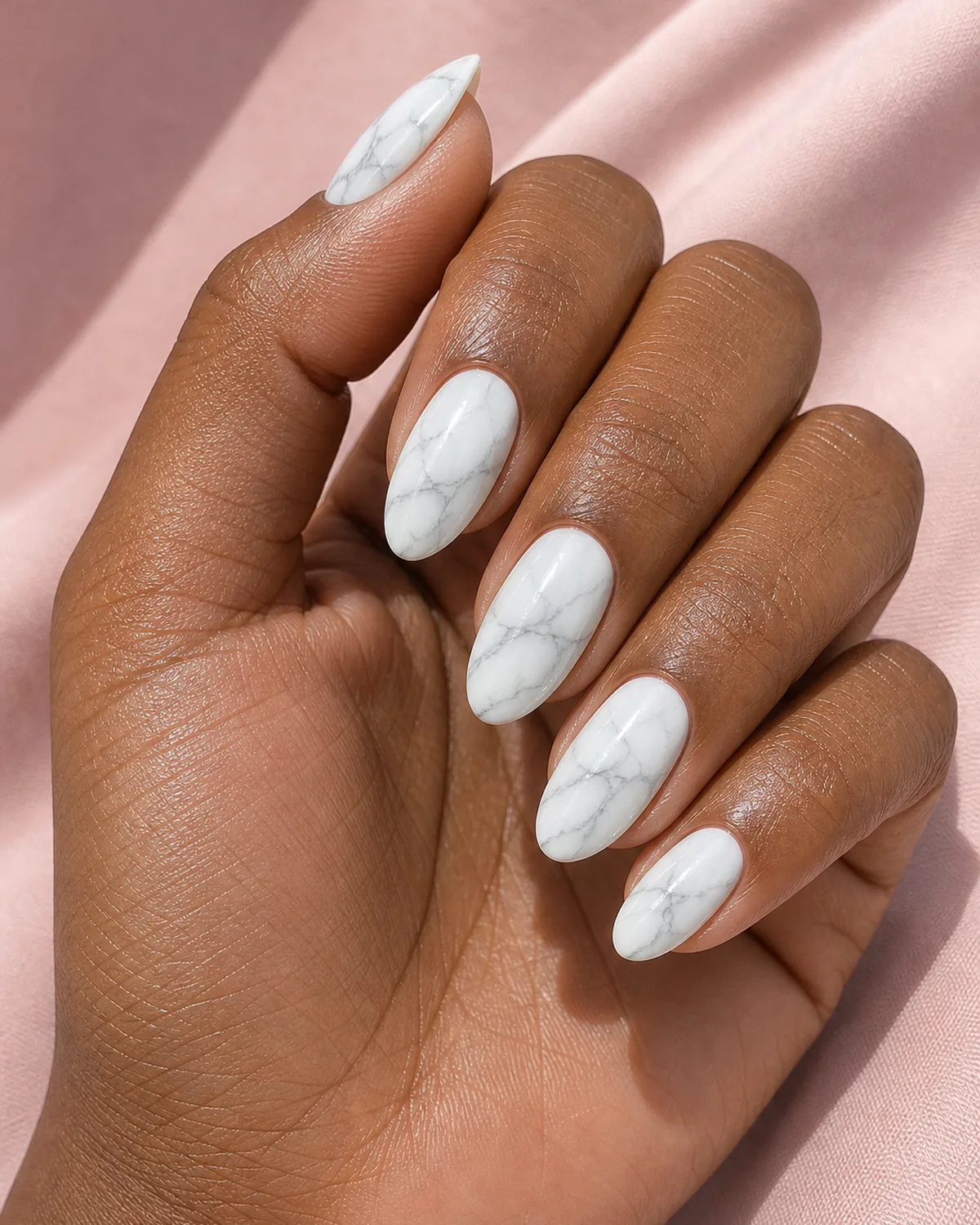 Close-up of medium-length almond nails in classic white-and-soft-grey marble with everyday editorial veining, on medium-deep skin against soft-pink fabric