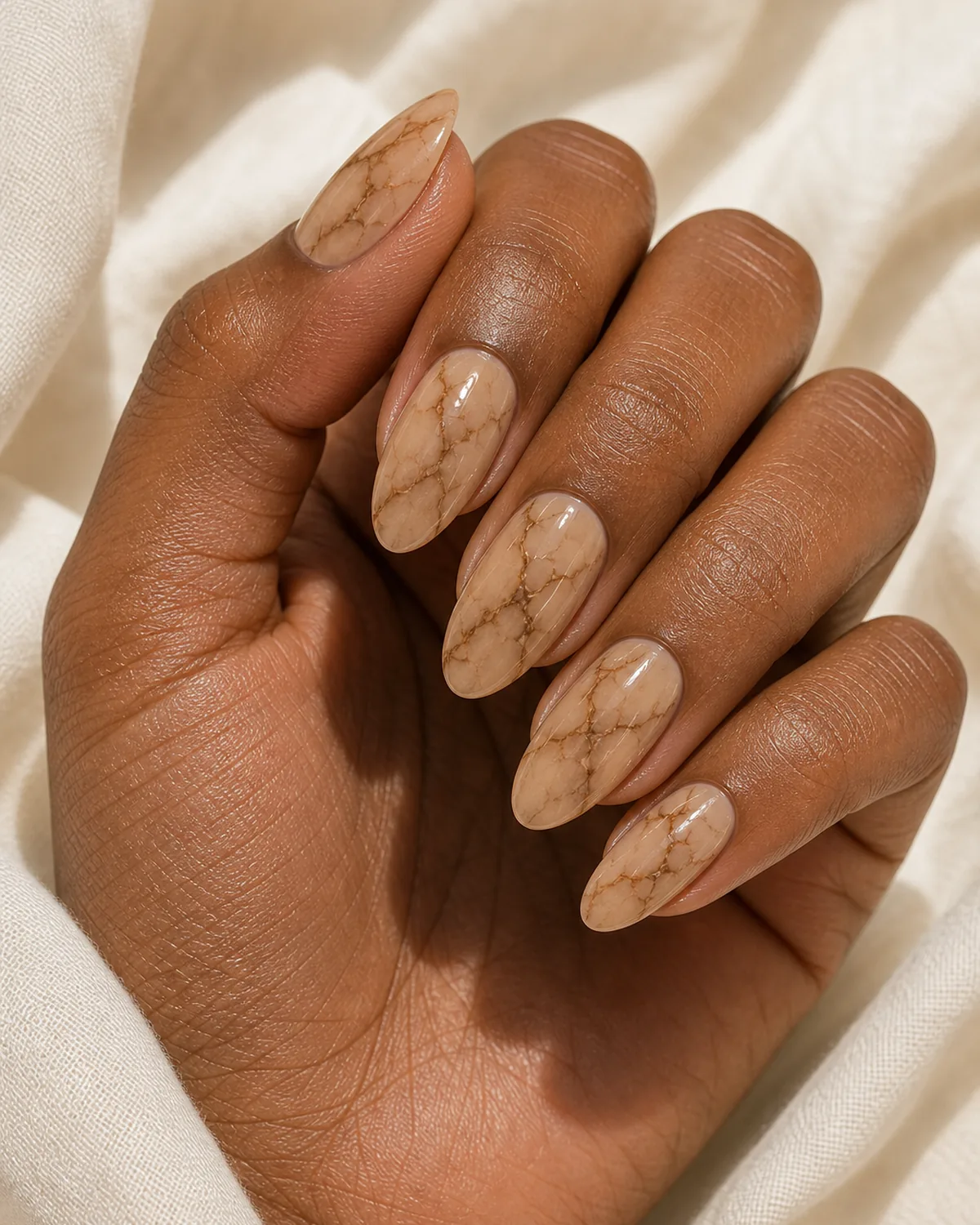 Close-up of medium almond nails in beige mocha marble with thin caramel-brown branching veins on a soft warm tan base, on deep skin resting on cream fabric