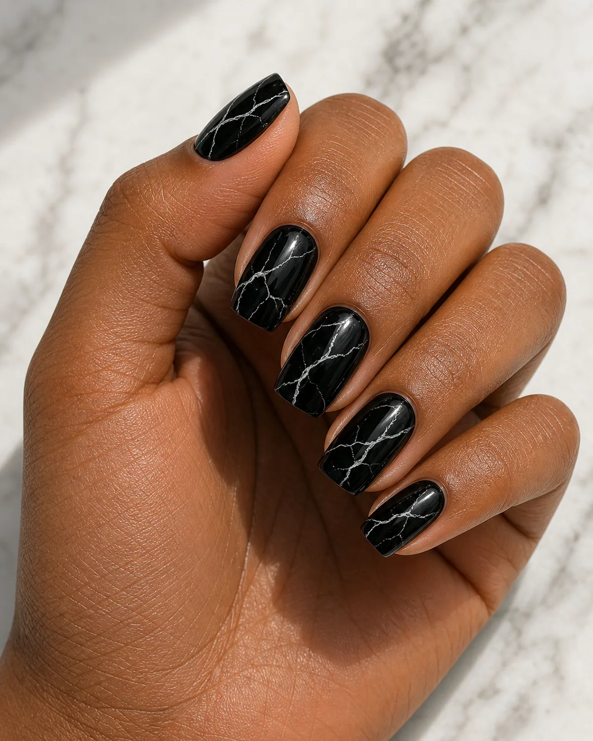 Close-up of medium squoval nails in black onyx marble with thin white branching veins on a glossy black base, on medium-deep skin resting on pale marble