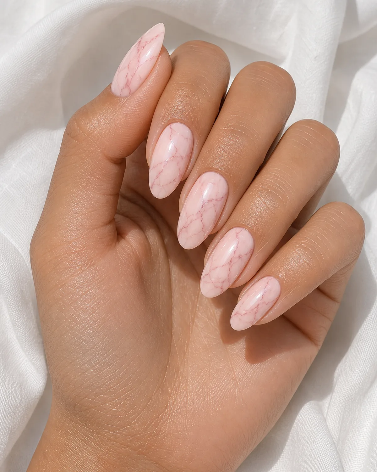 Close-up of oval nails in blush marble with thin rose-pink branching veins on a soft pink base, on medium skin resting on white fabric