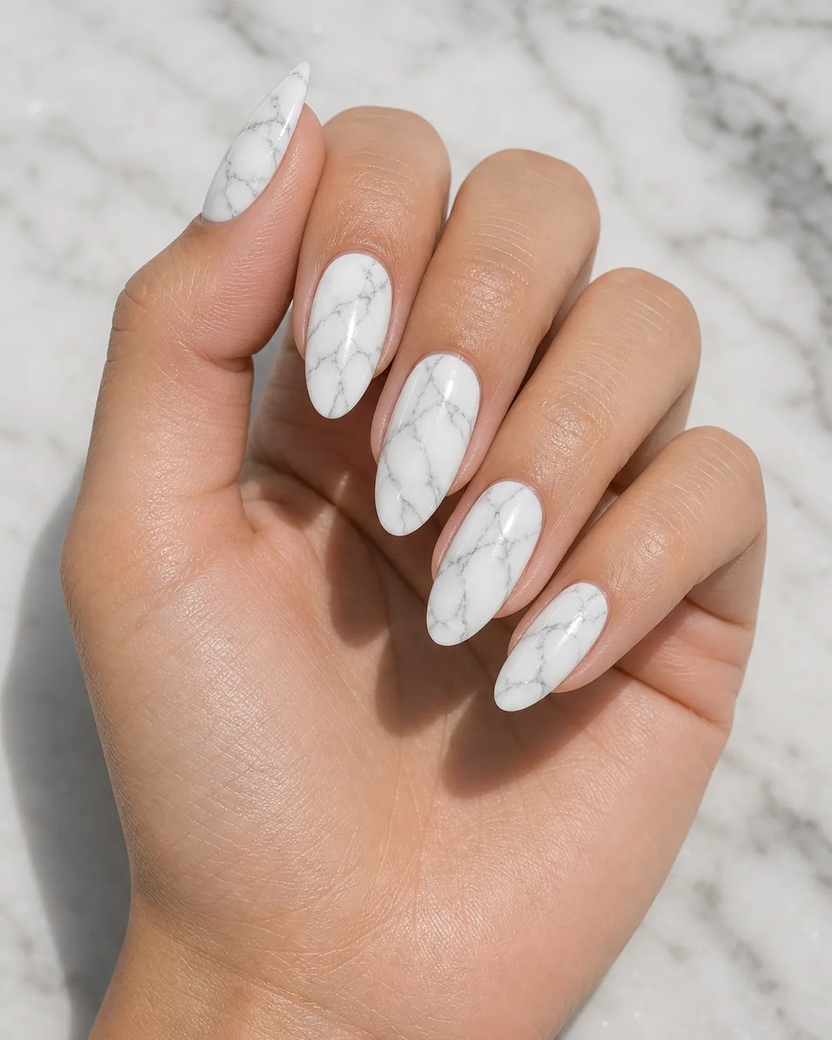 Close-up of medium almond nails in classic white Carrara marble with thin soft-grey branching veins on a milky white base, on light-medium skin resting on pale marble