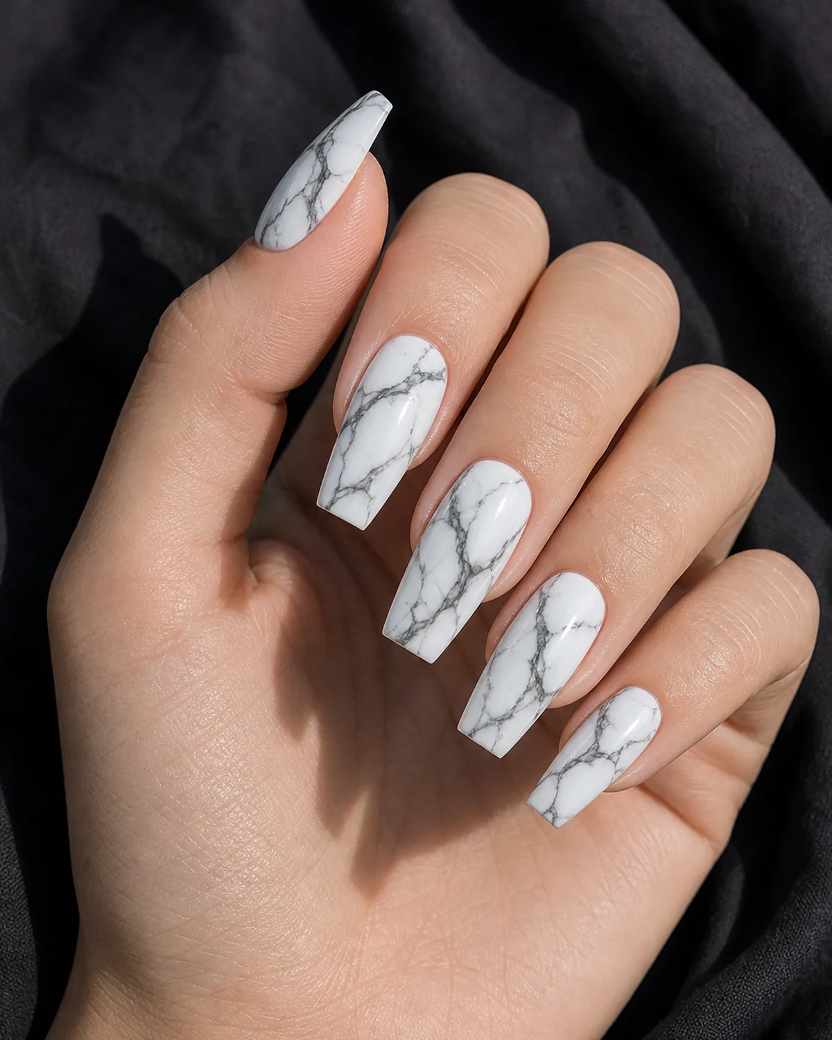 Close-up of coffin-shaped nails in statuario marble with bold charcoal branching veins on a bright white base, on fair skin against dark fabric