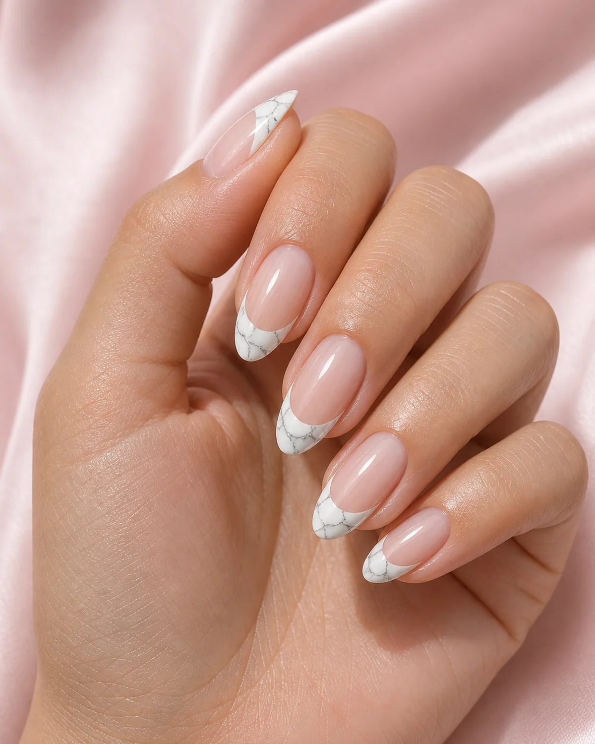 Close-up of almond nails in marble French tip with soft-grey veining hand-painted only inside a soft-white French smile, on light-medium skin resting on soft-pink silk