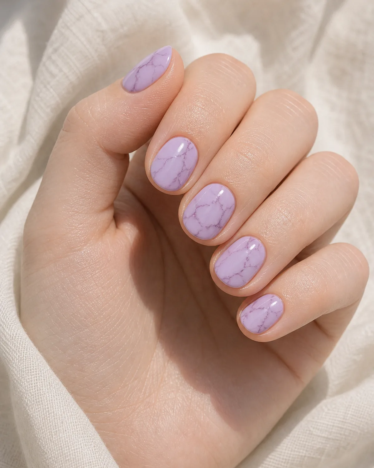 Close-up of short round nails in lilac marble with thin plum-purple branching veins on a pale lavender base, on fair skin resting on cream fabric