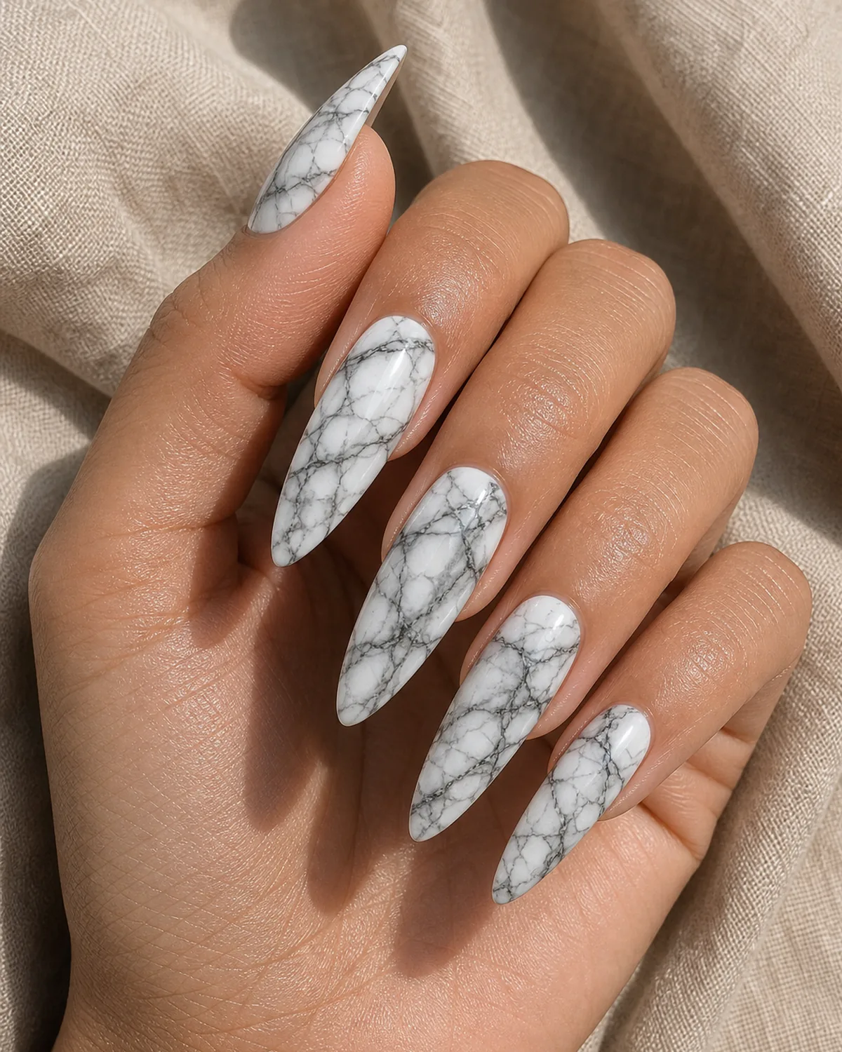 Close-up of long dramatic almond nails in maximum vein-drama marble with rich grey-and-charcoal branching veins on a milky white base, on medium skin resting on oat linen