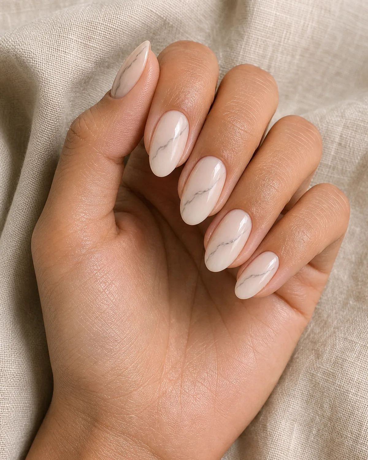 Close-up of short almond nails in one-vein minimal marble with a single thin grey diagonal vein on a sheer nude base, on tan skin resting on oat linen