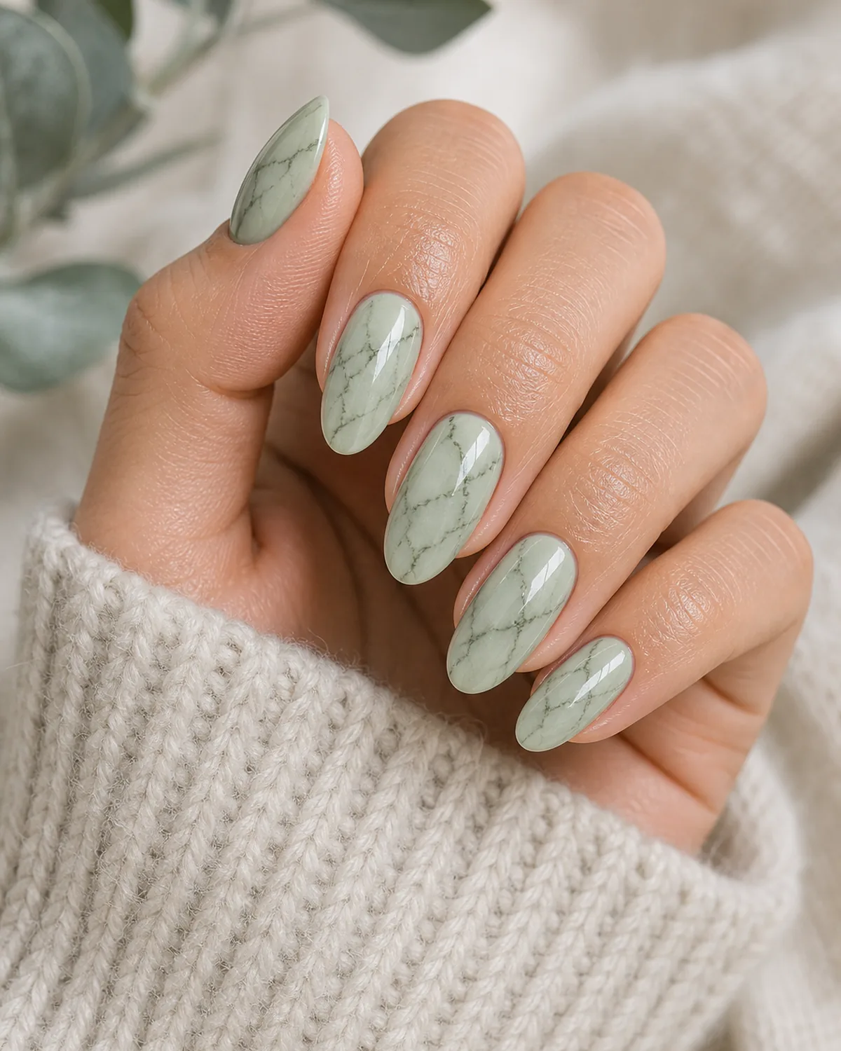 Close-up of medium oval nails in sage marble with thin mossy darker green branching veins on a pale sage base, on light-medium skin resting on a soft knit sleeve