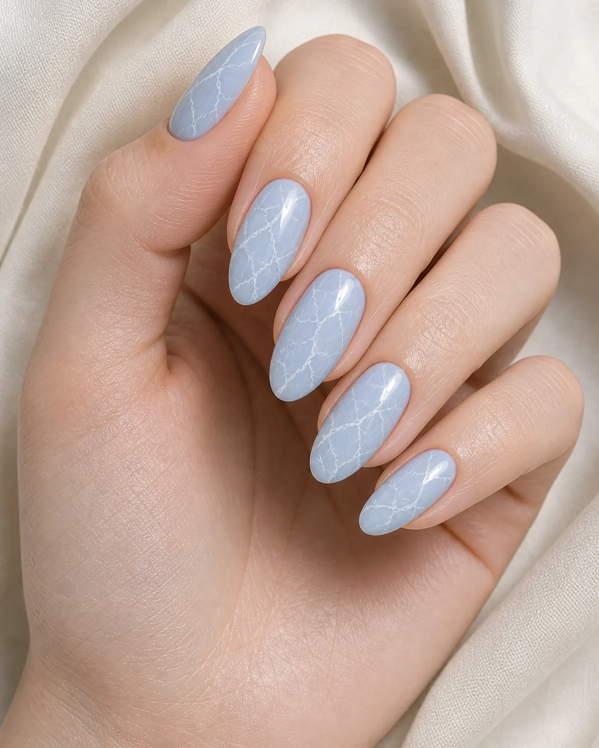 Close-up of almond nails in sky blue agate marble with thin white branching veins on a pale sky-blue base, on fair skin resting on cream fabric