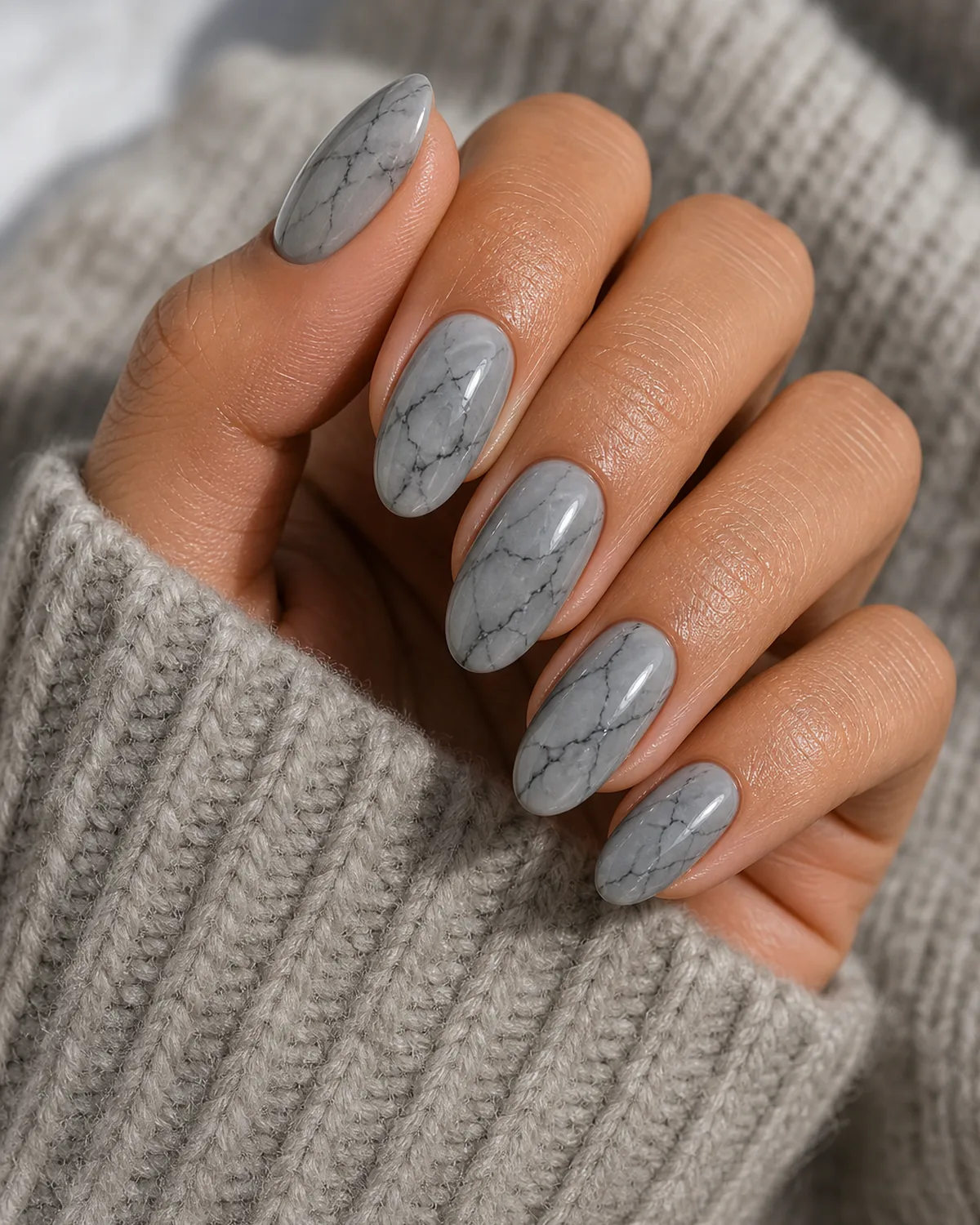 Close-up of medium oval nails in smoky grey marble with darker charcoal branching veins on a dove grey base, on tan skin resting on a soft knit sleeve