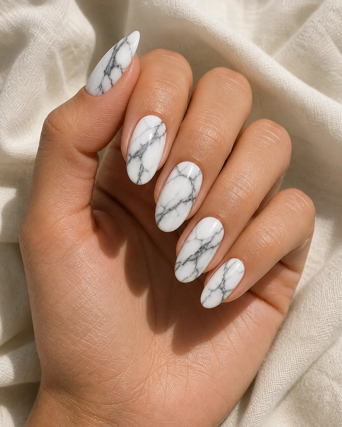 Close-up of medium oval nails in statuario marble with bold charcoal-grey branching veins on a bright white base, on medium skin resting on cream fabric