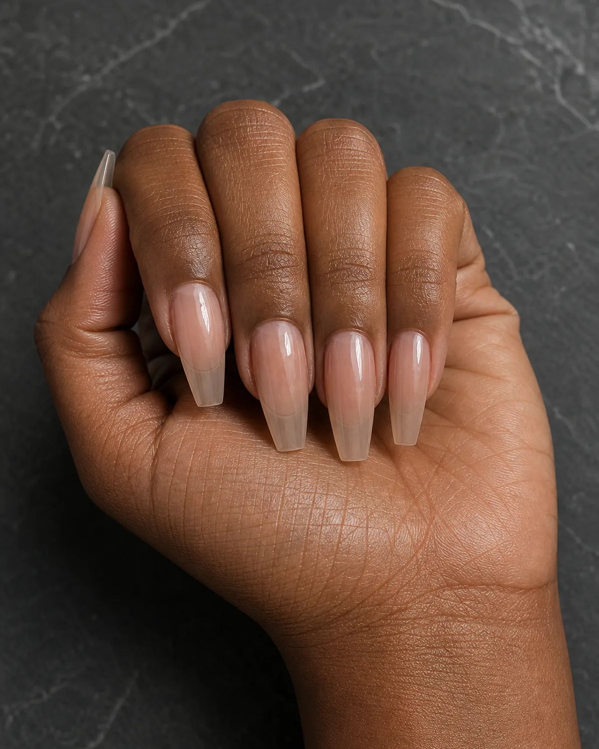 Close-up of long coffin nails in a bare-nail buffed polished look under a clear strengthener with high natural shine and no added color, on medium-deep skin closed into a soft fist on charcoal stone