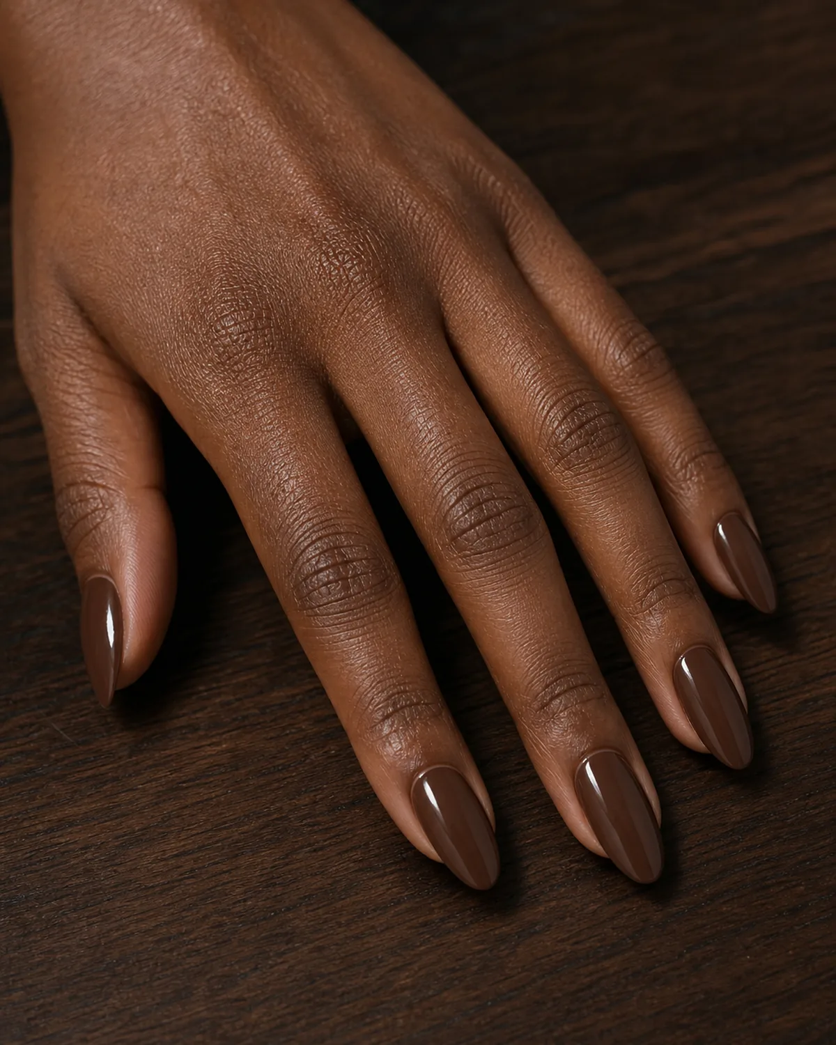 Close-up of medium almond nails in rich chocolate brown cream lacquer covering the entire nail surface in a smooth low-saturation finish, on deep skin against dark walnut wood