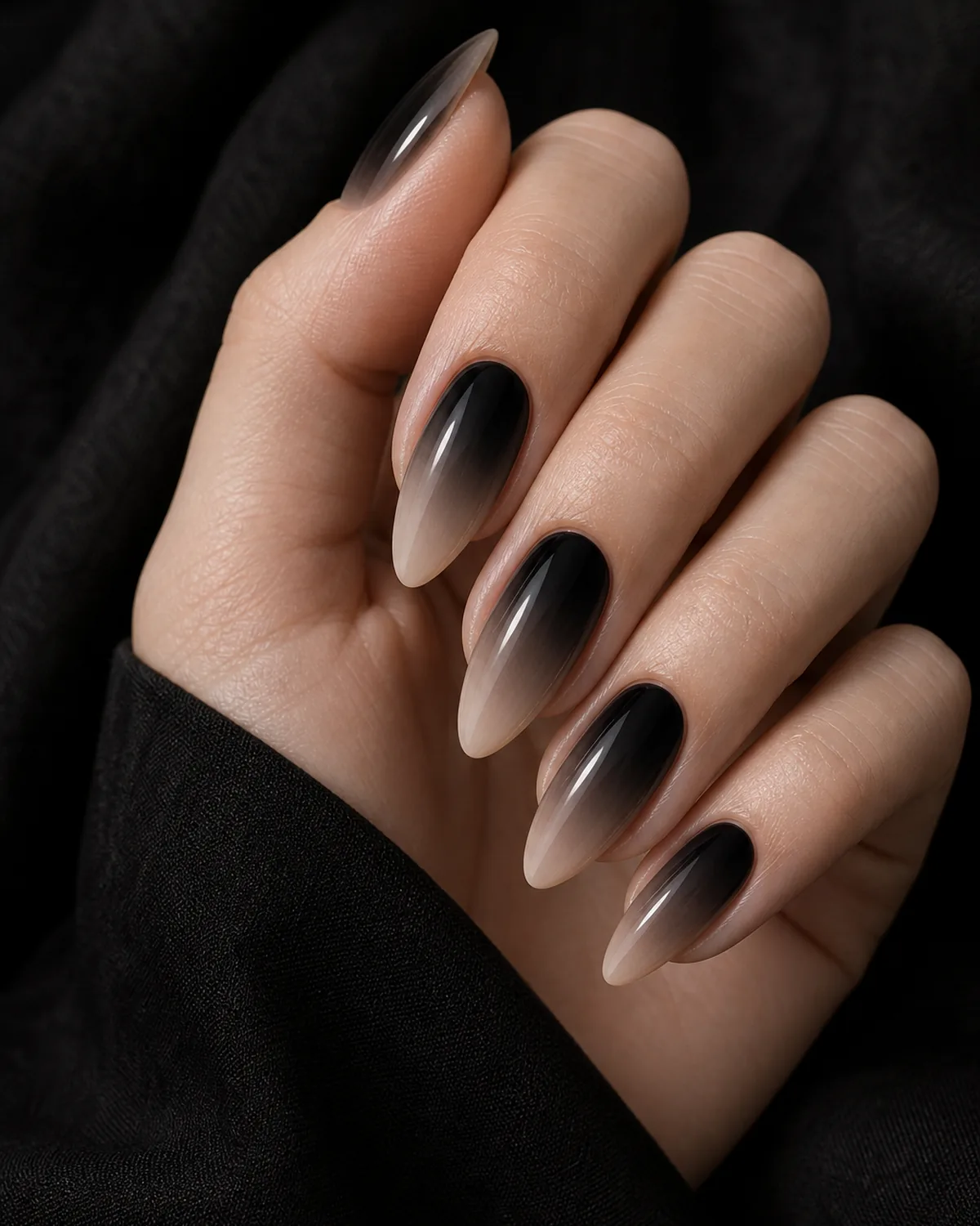 Close-up of long almond nails in a smoke ombre, true black fading dramatically into bare nude, edgy and minimal, on fair skin against dark fabric