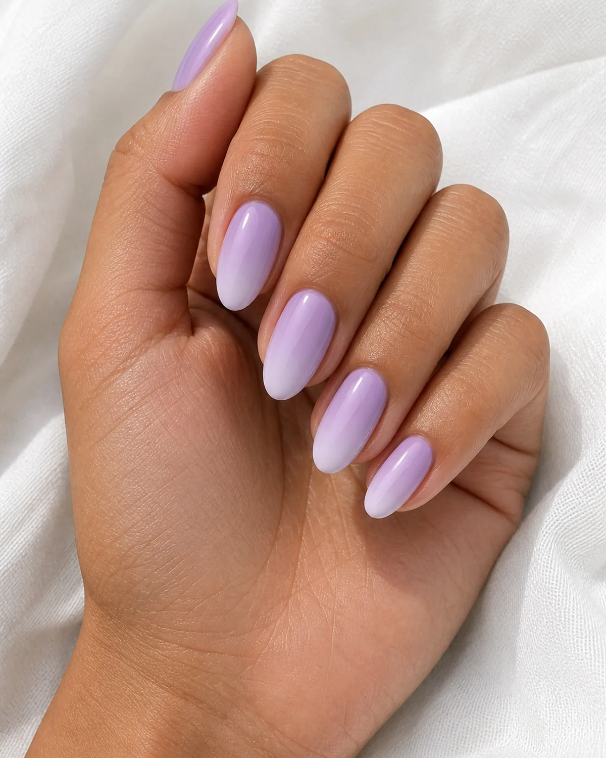 Close-up of oval nails in a lavender-to-lilac ombre, the blend pastel and seamless, on medium skin against white fabric