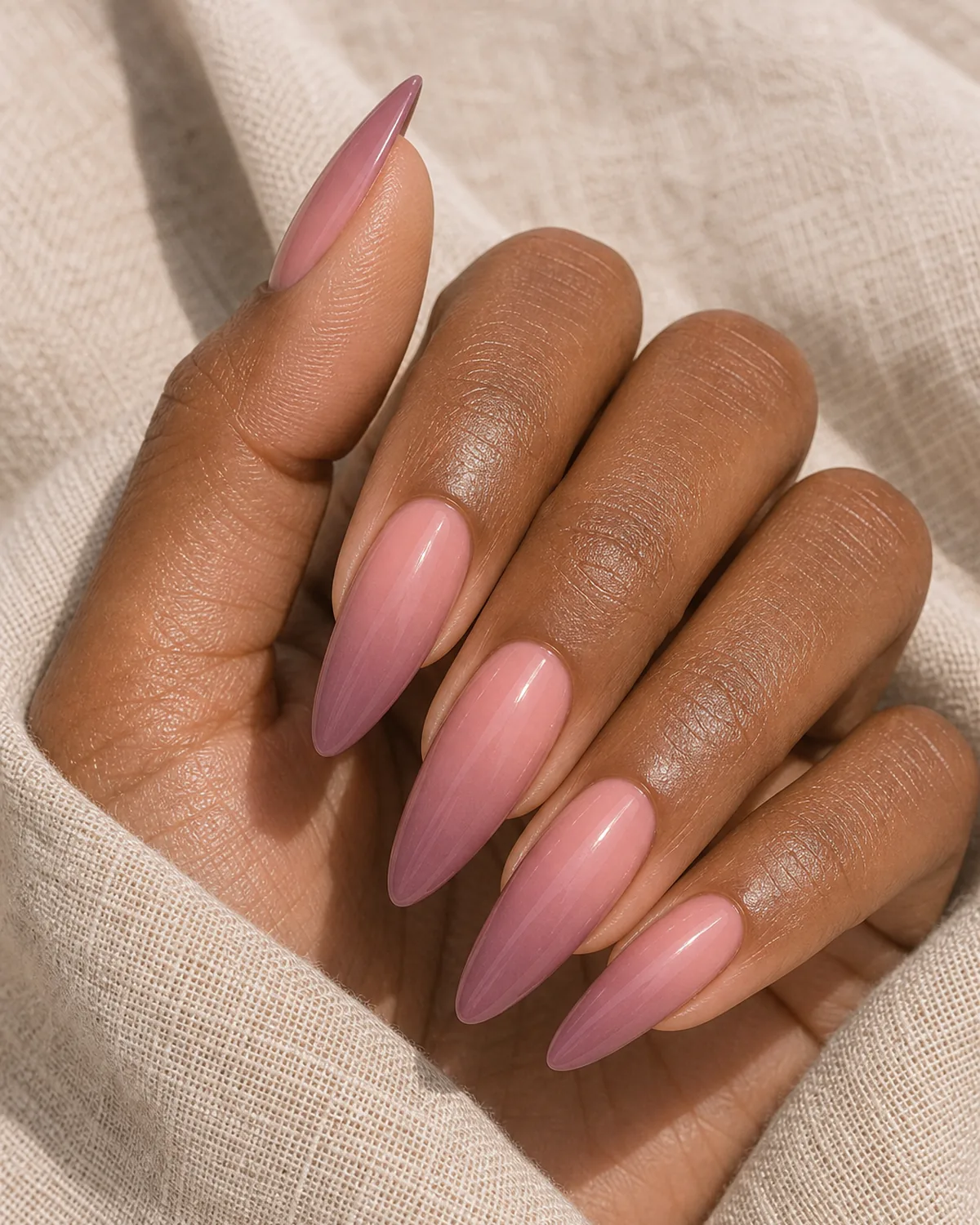 Close-up of long dramatic almond nails in a soft rose-to-mauve ombre, the gradient fading along the long taper, on medium-deep skin resting on oat linen