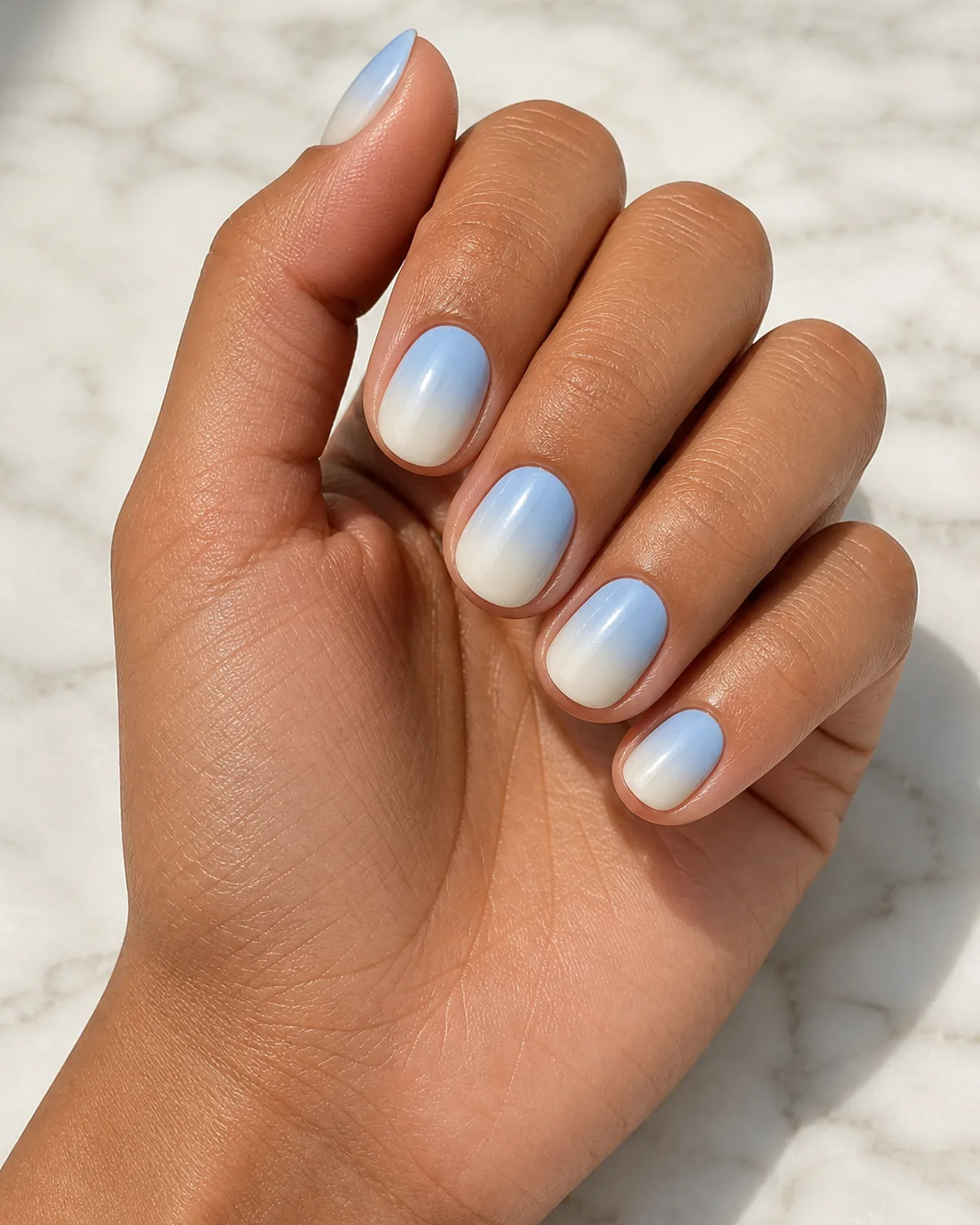 Close-up of short round nails in a sky-blue-to-cream ombre, the blend cool and airy, on tan skin resting on pale marble