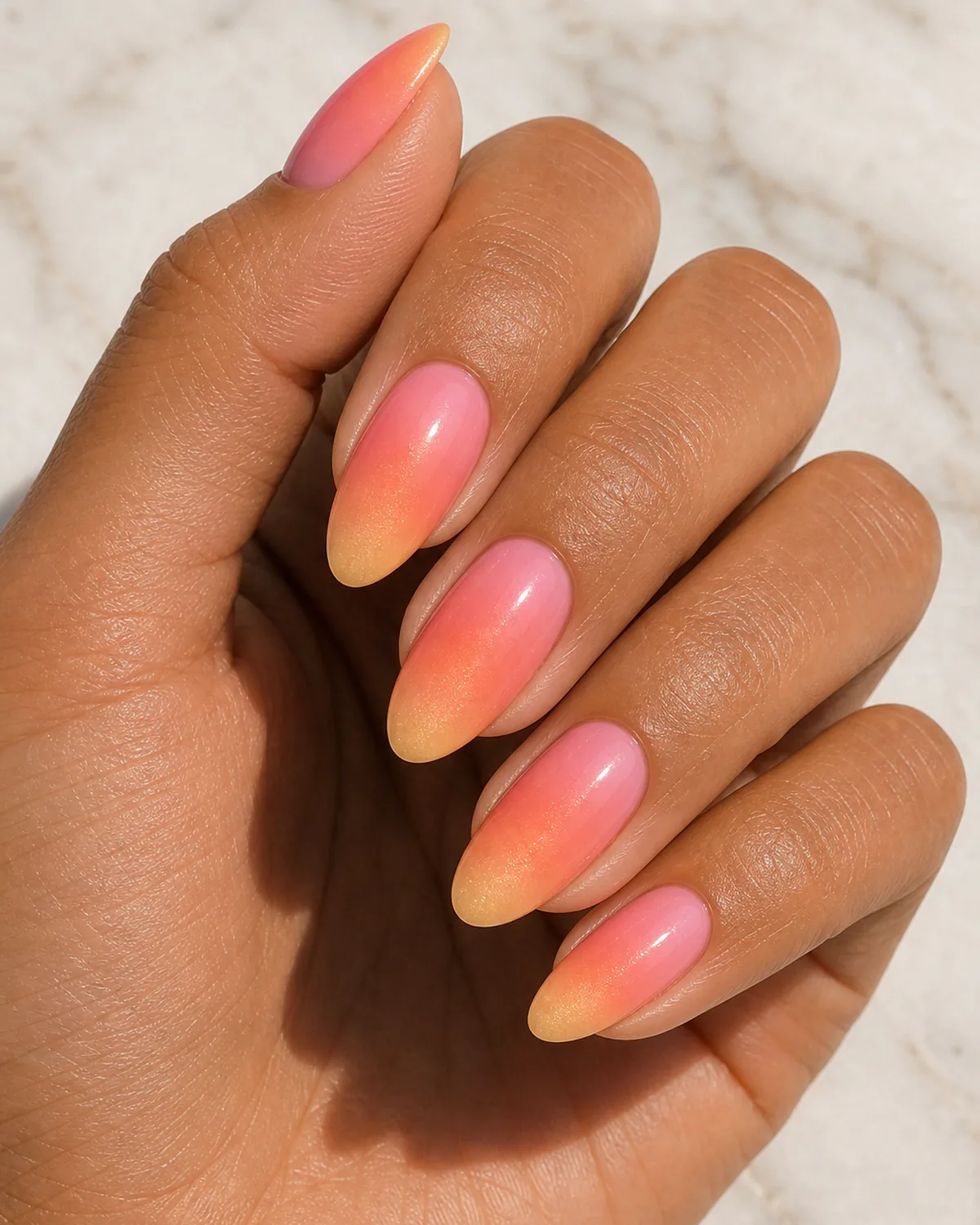Close-up of medium almond nails in a sunset three-tone ombre fading pink into coral into warm gold, on medium skin against pale marble